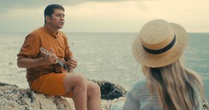 Couple enjoying holidays sitting on sea beach with ukulele under sunset sky in evening time. Man playing, woman listening and sing music, relaxing , travel day. 4K video