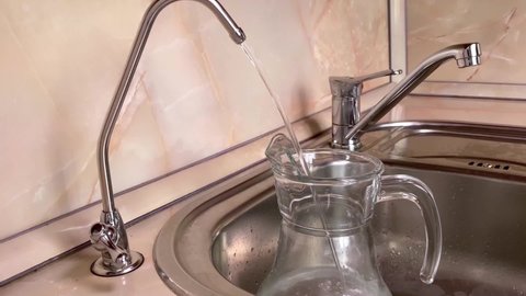 Pouring filtered water into glass pitcher from water filter. Close up of jug, sink and faucet. Drinkable water in kitchen