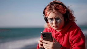 preteen girl is viewing learning video in smartphone, listening to sound by headphones on sea beach