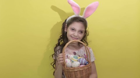 4k. Happy easter. Beautiful cute little girl in Easter bunny ears holds a basket with eggs on a yellow background. Easter activities for children. Hello spring and easter concept