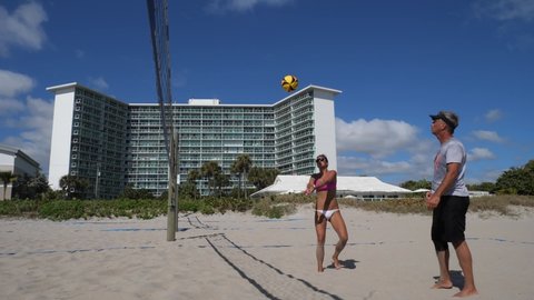 Deerfield Beach, Florida  USA - February 26 2022:  Guys and girls play coed volleyball in slow-motion in the sand beneath the net in the late-morning sun with condominiums in the background.