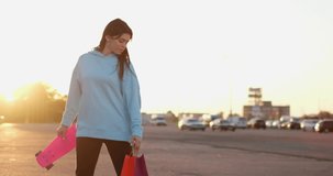 Young brunette woman spins around holding skateboard and colorful bag with happy smile. Cars drive on parking lot near shopping center at sunset