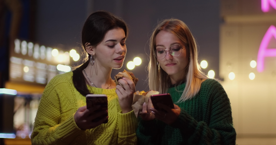 Women discuss news scrolling through smartphones and eat hot dogs in paper wrapping. Busy street with garlands and neon signs illuminating at night closeup