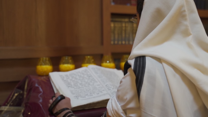 A Jew prays in the synagogue. A man stands near the presidium and reads a book. Back view
