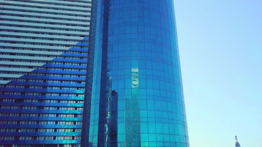 A close-up shot of the reflections of the sun, clouds and sky on the glass windows of a city skyscraper. The drone flies up, a close-up of the floors of building. Blue color. Real estate and hotel.