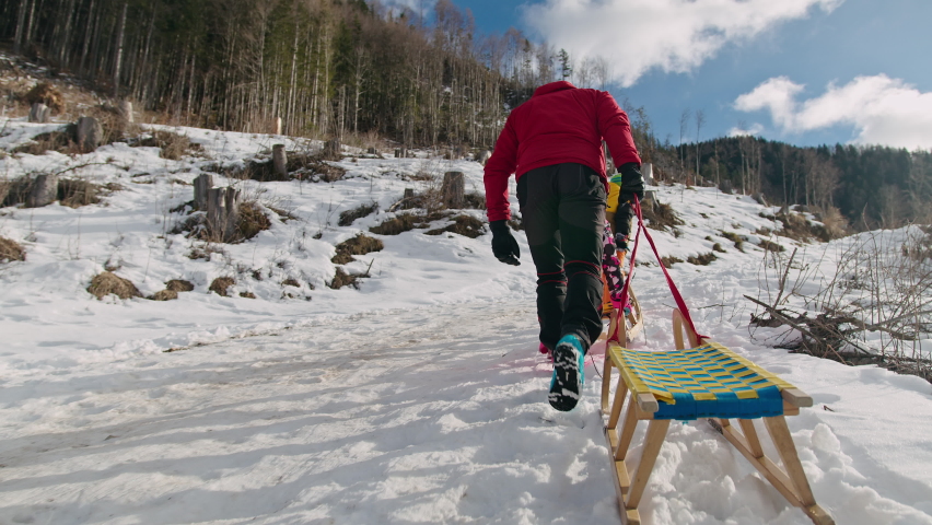 Father and kids, boy and girl, pulling empty wooden sleds uphill, walking through snow one after the other, tracking rear view.