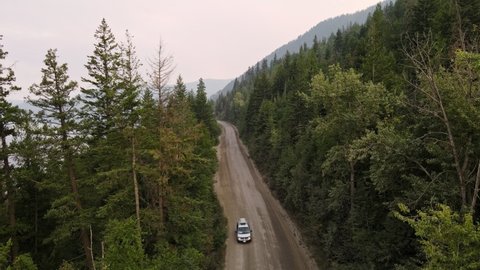 Drone footage of a car driving through a dry boreal forest in British Columbia at sunset. Aerial opposite fly-over shot