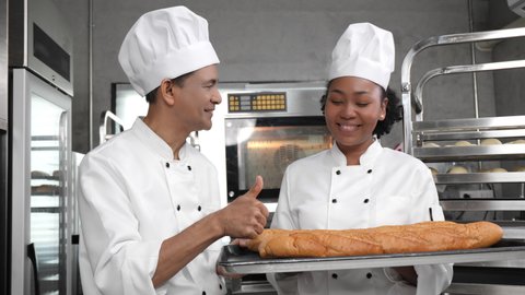 A senior Asian male baker giving thumbs up to a young African female trainee baker holding a tray with freshly baked French breads, compliment on her perfect loaves of bread at a bakery kitchen.