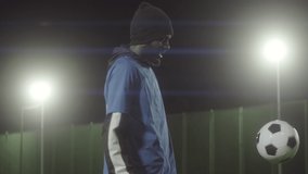 Medium portrait of fit African American male athlete wearing uniform and warm hat, practicing soccer moves with ball, then posing on camera standing on football field in spotlight at night - Powered by Shutterstock - Get 15% off with code: PIKWIZARD15