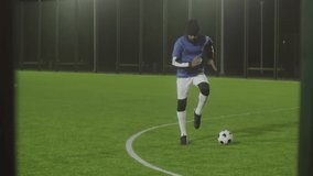 Long shot of young African American male athlete wearing uniform, warming up on football field in spotlight at night, soccer ball on grass - Powered by Shutterstock - Get 15% off with code: PIKWIZARD15