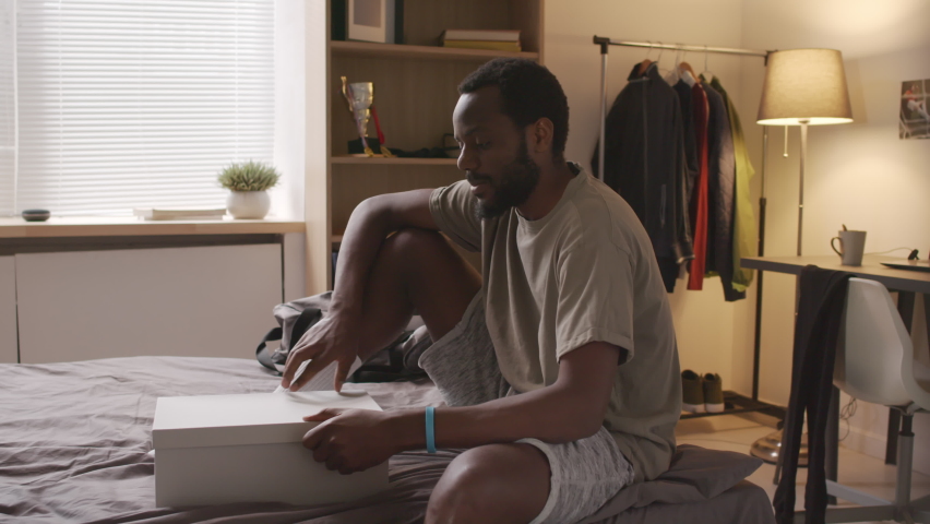 Medium long of young Black man sitting on bed at home, unpacking shoe box with new soccer boots at daytime