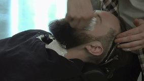 Barber prepares young hipster customer for hot shave in salon chair