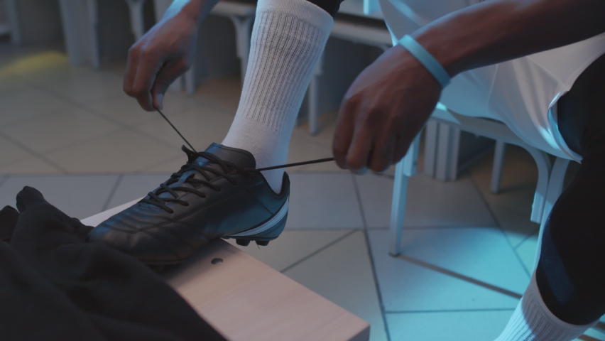 Tilting up of young African American male athlete sitting in locker room, tying up laces on cleats early in morning - Powered by Shutterstock - Get 15% off with code: PIKWIZARD15