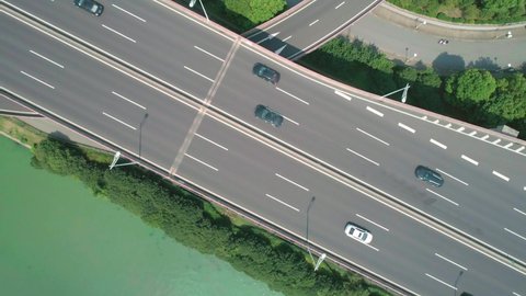 Aerial top down view of a highway overpass multilevel junction with fast moving cars surrounded by green trees and with a river on a side on a sunny day.