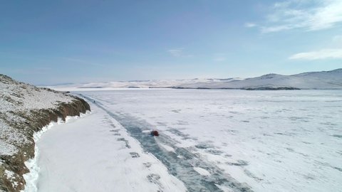 Aerial view on the hovercraft driving on cracked snowy ice of Baikal along the rocky cliff. Drone follows the vehicle. Beautiful winter landscape of lake Baikal