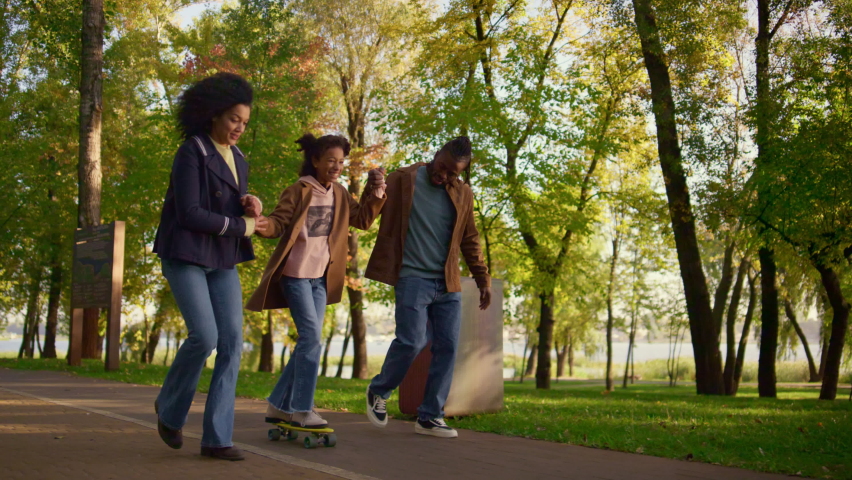 Parents teaching girl riding longboard in park. Supportive family encourage kid. Joyful african american couple holding daughter hands help balancing. Loving father caring mother enjoying parenthood. Royalty-Free Stock Footage #1087874885