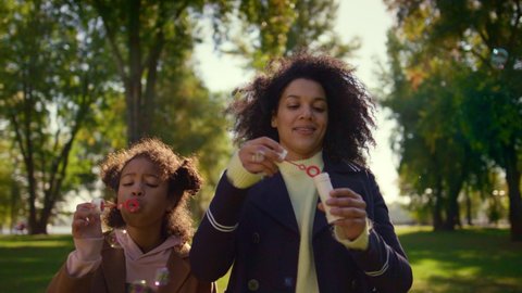 Parent kid blowing soap colorful bubbles in green sunny park together portrait. Smiling african american mother playing enjoy warm autumn day with cute daughter. Parenthood emotional bonding concept.