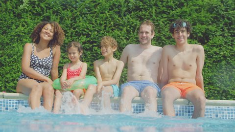 Portrait of multi-racial family relaxing sitting on edge of swimming pool on summer vacation shot from underwater as camera break the surface - shot in slow motion