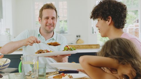 Father serving as multi-racial family sitting around table in kitchen at home eating meal together - shot in slow motion