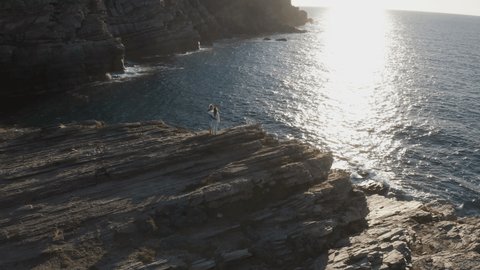 Couple on top of cliff above the ocean enjoy stunning sunset from above. Drone aerial view 