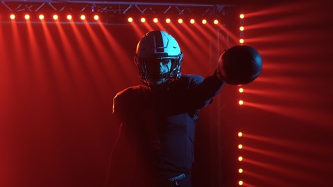 American football player in helmet and uniform enters the playing stadium. Confident agressive player challenge opponents while standing in dark against red backlight and smoke. Close up. Slow motion.