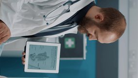 Vertical video: Male doctor showing human skeleton illustration on digital tablet to senior woman at checkup examination. Physician and patient looking at osteopathy diagnosis with bones and spinal
