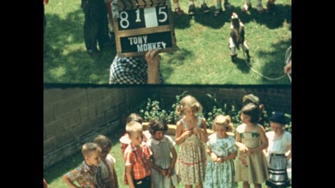 1950s: Young girls and boys stand around a grassy lawn opposite a monkey in clothing. A blonde boy stoops and reaches out, the monkey comes over then runs back. Girls squat and reach for the monkey