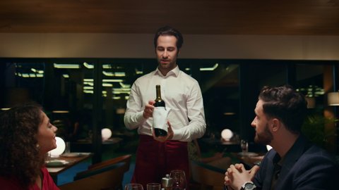 Restaurant waiter pouring wine serving mixed race couple at fancy cafe table. Two romantic partners drinking alcohol glass in modern style hotel bar. Lovers enjoy dating indoors. Luxury dining concept