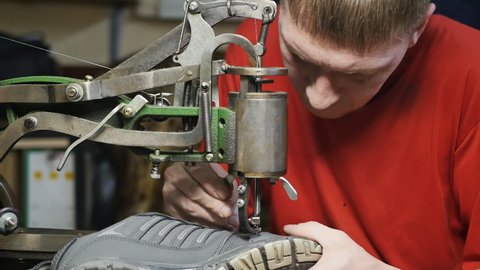 Caucasoid male shoemaker stitches the seam of a sneaker with a new thread using a retro machine with a manual drive and fully mechanical