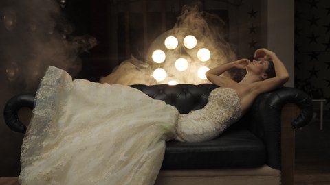 Young woman in wedding dress poses lying on leather sofa