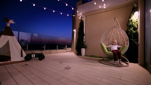 kid is relaxing in hanging chair, having fun on rooftop patio at summer evening