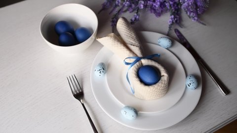 Festive decorated Easter table. Blue egg with a napkin like an Easter bunny and blue flowers on a white table. Holiday, chocolate eggs, white plates. Painted eggs.