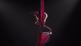 Young female circus gymnast spins on aerial silk and demonstrates stretching. Difficult acrobatic stunts at height on black background with backlight. Video for sports, acrobatic, circus school. Slow.