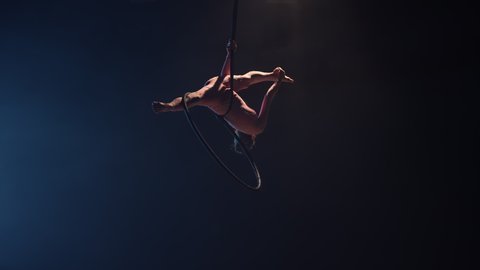 A young woman gymnast performs acrobatic stunts on an air hoop under the dome of a circus. Exciting acrobatic show in a dark smoky studio with blue backlight. Slow motion.