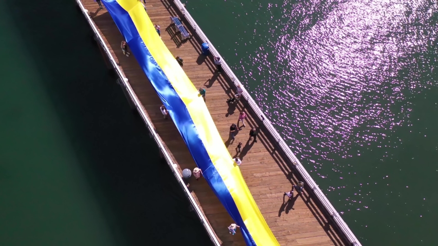 CALIFORNIA - CIRCA 2022 - Aerial Ukranian protesters with large Ukraine flag protest the Russian invasion of their country, on the Malibu pier. Royalty-Free Stock Footage #1087896125
