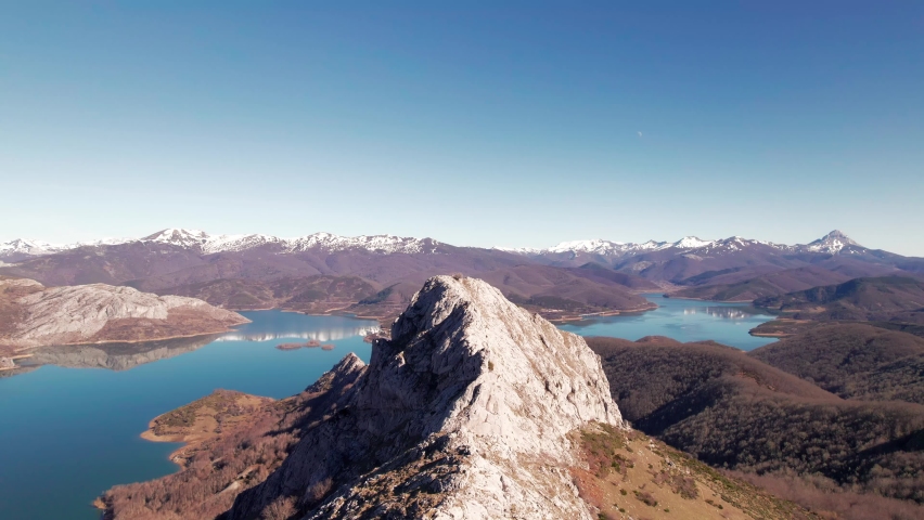 Aerial shot of Pico Gilbo in León, Spain. Big mountain peak on the center of the screen. Snowy mountain range next to a lake below a a clear blue sky with no clouds.