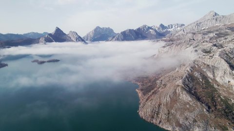Aerial view, drone above clouds and a huge mountain range in León, Spain. Misty, foggy, cloudy winter day over a massive lake. Snowy mountain summit far away, long distance. Dark blue water.