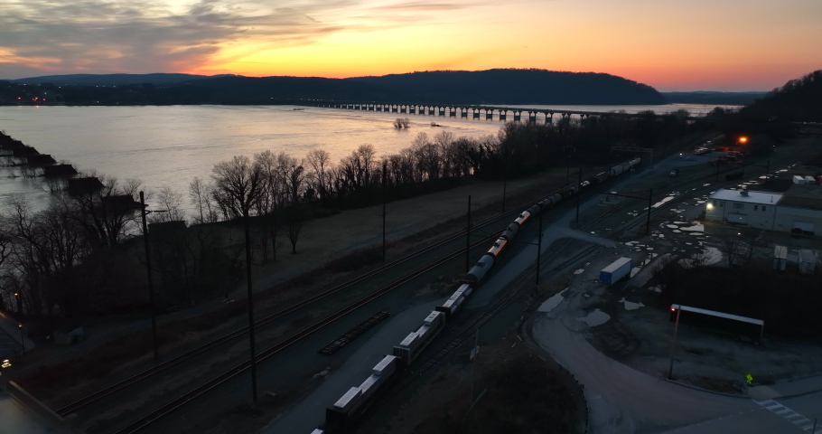Railroad train at night travels on tracks beside river. Evening sunset view. Transportation freight theme.