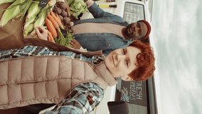 Vertical shot of young Caucasian woman and African American man smiling, waving and talking on camera via online video call while selling fresh vegetables from farm at outdoor market