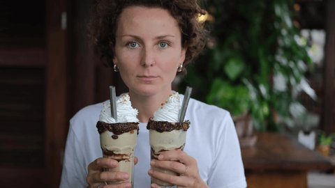 Funny curly young woman playing, enjoying, eating whipped cream from two glasses of chocolate milk drinks