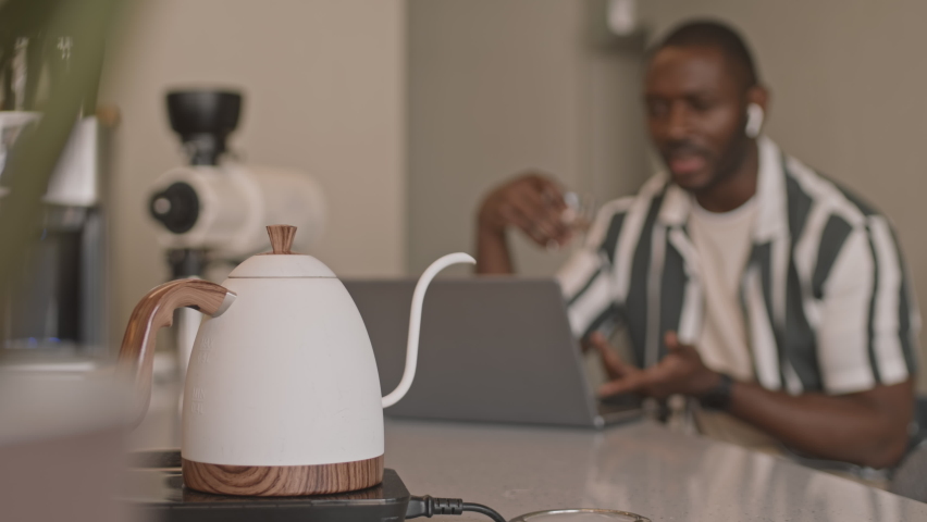 Close-up of white retro-style kettle on counter bar of coffee shop in selective focus and young African American man having video chat on laptop in blurred background