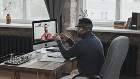 Middle eastern man in wireless earphones sitting at desk in loft office and speaking with business partner on web call on computer while having online meeting