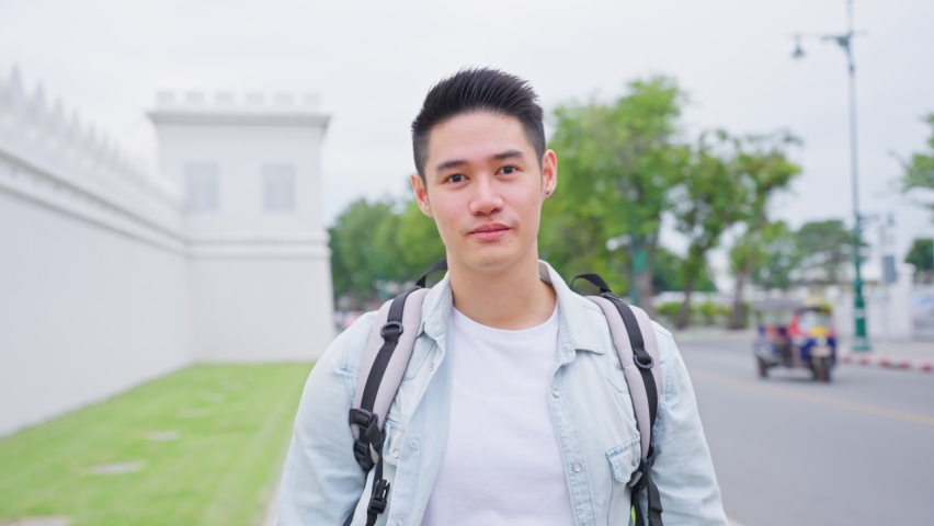 Asian attractive man backpacker walking around beautiful road in city. Portrait of handsome guy tourist traveler travel alone on street, enjoy spend time on holiday vacation trip and look at camera.