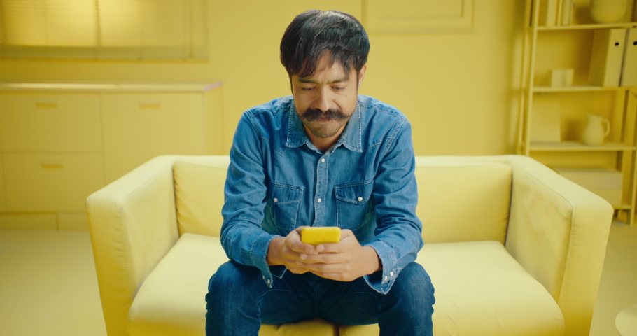 Portrait of Hispanic man with mustache looking at the mobile phone screen and looks surprised with mouth open while sitting on the sofa at home. Shocked man laughing at social media on cellphone