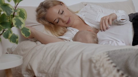 Young smiling mother strokling her sleeping son. Concept of breastfeeding, maternity and family togetherness. Woman laying on bed and feeding with breast her baby boy