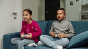 Multiethnic preteen young friends competing by playing video game while sitting on sofa indoors. Cheerful white girl and cute black boy spending leisure time together, having fun at home. Moving shot