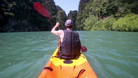 Man sails on yellow kayak on azure water in canyon rowing with pink paddles. Tourist enjoys beautiful view with lush trees in Thailand backside view