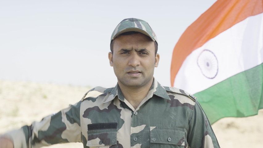 army officer saluting by looking at camera in front of waving Indian flag - conept of patriotism, respect and pride.