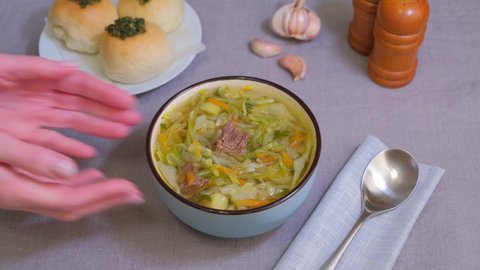 Shchi with pampushki (buns) and lard. Soup of cabbage, potatoes and meat. Traditional Russian and Ukrainian cuisine. Woman puts a bowl of soup on the table. Close-up.