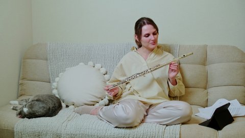Happy woman musician with flute looks into tablet at home on sofa in living room